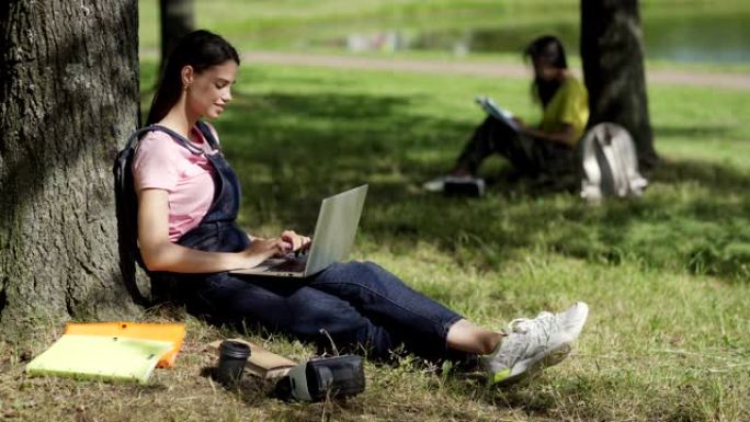 女学生坐在树下并在公园的笔记本电脑上打字的侧视图。创意女孩抬头思考，课本，咖啡杯和附近的VR眼镜。背