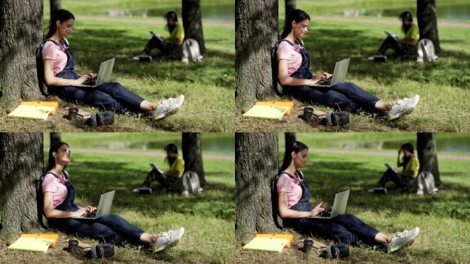 女学生坐在树下并在公园的笔记本电脑上打字的侧视图。创意女孩抬头思考，课本，咖啡杯和附近的VR眼镜。背