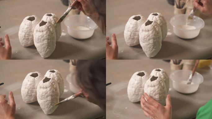 一个专业的女陶工手持刷子，用液体物质覆盖现代风格的陶瓷雕塑的前视图。创造陶瓷产品概念