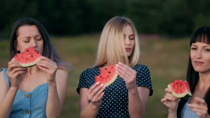 日落时，三个穿着蓝色连衣裙的年轻迷人女性朋友正在吃西瓜和微笑。