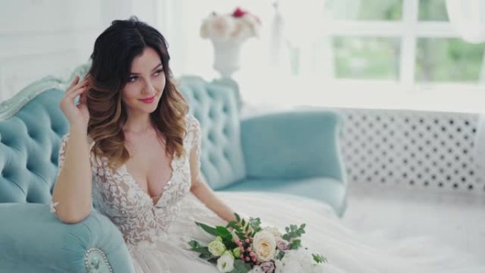 穿着婚纱的魅力新娘坐在家里带花的沙发上。