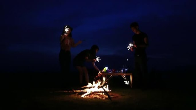 一群亚洲朋友的游客正在弹吉他，而他放松的朋友正在一起坐在帐篷附近的篝火旁听，扔柴火和玩烟火