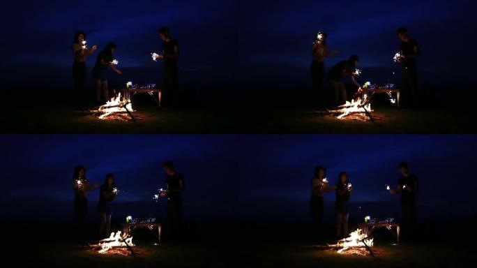 一群亚洲朋友的游客正在弹吉他，而他放松的朋友正在一起坐在帐篷附近的篝火旁听，扔柴火和玩烟火