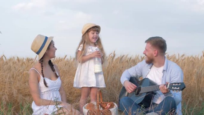 家庭在野外度假，年轻的父亲弹吉他，与妻子在女儿喝酒时大笑，并在小麦谷物作物草甸的户外野餐中舔牛奶胡须