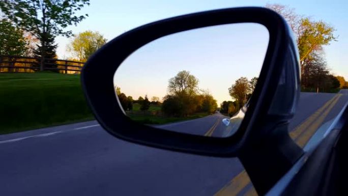 日落时驾驶侧镜的乡村道路视图。司机的视点POV沿着美丽的乡村街道在黄昏附近俯视侧视镜。