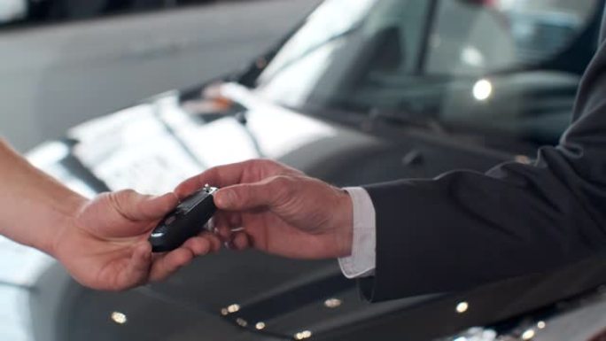 年轻人把车钥匙给买家。男人在买了车的背景下在漂亮的汽车经销店握手。