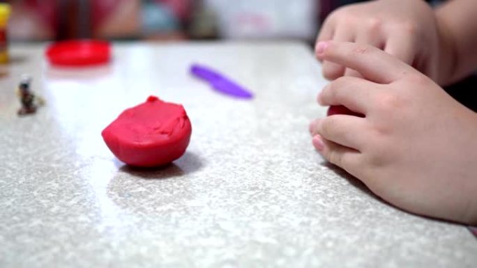 一个在明亮的房间里玩粘土的孩子。近距离接触。她手里拿着红粘土。用紫色塑料刀。孩子的身体在右边。