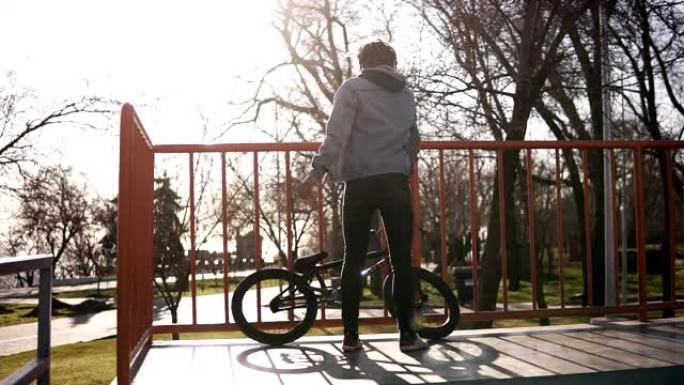 一个留着辫子的男孩坐在constration上，然后在阳光明媚的日子开始在滑板公园里骑BMX自行车技