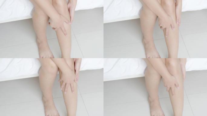 特写镜头美丽的年轻亚洲女人坐在床上抚摸着柔软光滑的皮肤在卧室里的腿，女孩应用润肤霜和乳液与治疗护理，