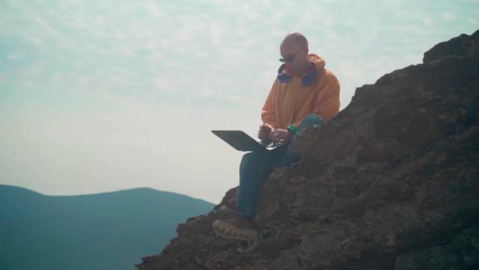 一名身穿黄色夹克、蓝色牛仔裤、戴着眼镜的男子坐在悬崖边，正在用笔记本电脑工作。