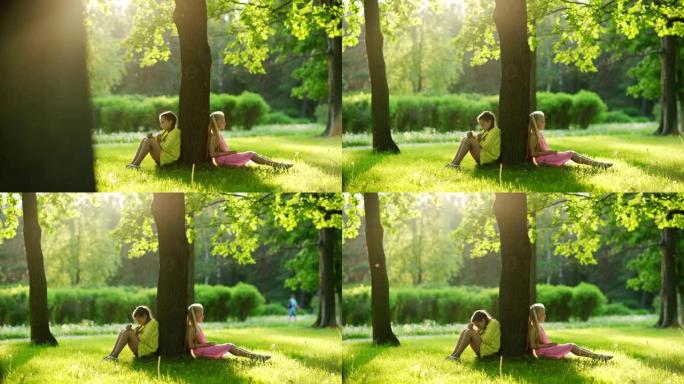 追踪两个不幸的小女孩坐在公园的草地上，背靠背靠在树干上，在阳光明媚的夏日里最好的朋友发生冲突后生闷气