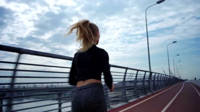 后视图跟踪在多云天空下在桥路自行车道上跑步的运动服年轻女子的右镜头