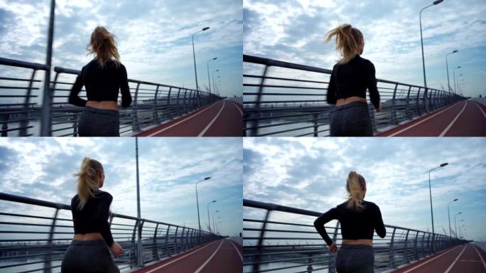 后视图跟踪在多云天空下在桥路自行车道上跑步的运动服年轻女子的右镜头