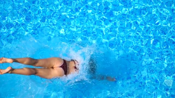 年轻女子跳进游泳池，溅出清澈的水的俯视图。晴天，面目全非的家伙在水盆里游泳。男人在暑假放松。慢动作特