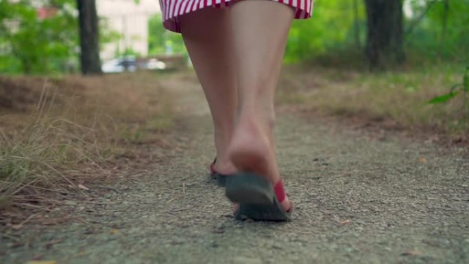 女性的腿在公园的人行道上行走。一个穿着凉鞋和条纹连衣裙的女孩走路。女性腿的特写。