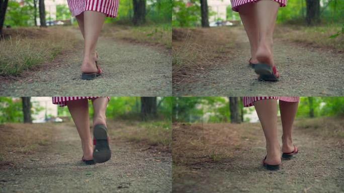 女性的腿在公园的人行道上行走。一个穿着凉鞋和条纹连衣裙的女孩走路。女性腿的特写。