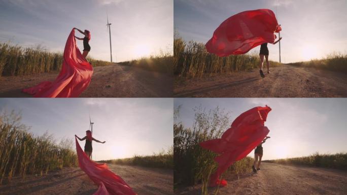 女人带着一块红布跑到野外的风力发电机上。日落时的风车。慢动作。时尚的时髦女人