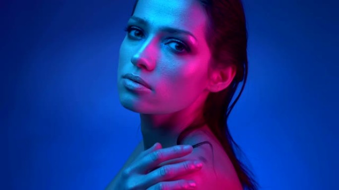 发光的模特在蓝色和粉红色的霓虹灯中闪烁着闪亮的妆容，看着相机温柔地抚摸着她的肩膀。