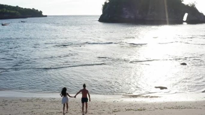 男人和女人在日落时沿着热带海滩散步，跟踪年轻夫妇在海滩上玩耍的航拍。日落时分，夫妇在海滩边的空荡荡的