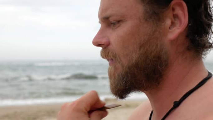 一位在大海背景上留着胡须的年轻人正在用木梳子梳理胡须。4 k视频