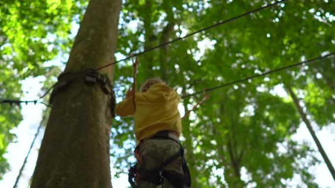 穿着安全带的小男孩在森林冒险公园的一条路线上攀爬。他爬上高高的绳索小径。户外娱乐中心，为成人和儿童提