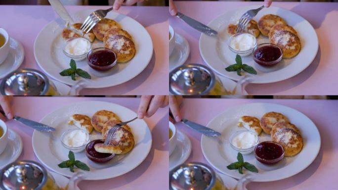 女性的手在餐厅用刀叉切了美味的奶酪煎饼。一个女孩在吃果酱和酸奶油的奶酪煎饼。