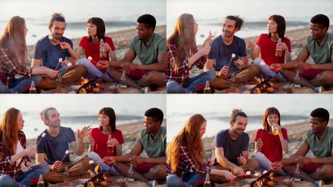 四个轻松多样的朋友坐在海滩篝火旁，聊天，烧烤，吃棉花糖和喝啤酒，跟踪左中枪