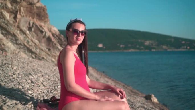 穿着红色连体泳衣和眼镜的美丽怀孕女孩坐在海滩上。