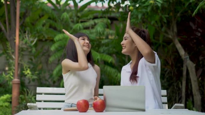 两名快乐的年轻亚洲女性使用笔记本电脑工作或学习，拍手成功，击掌五，在花园里一起拳头。庆祝目标获胜者。