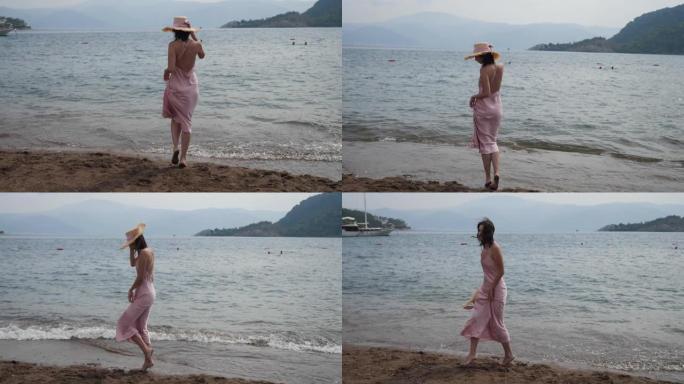 穿着粉色丝绸连衣裙和草帽走在沙滩上的女人