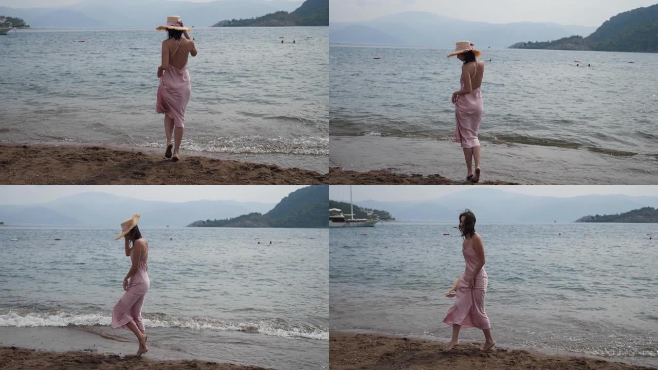 穿着粉色丝绸连衣裙和草帽走在沙滩上的女人