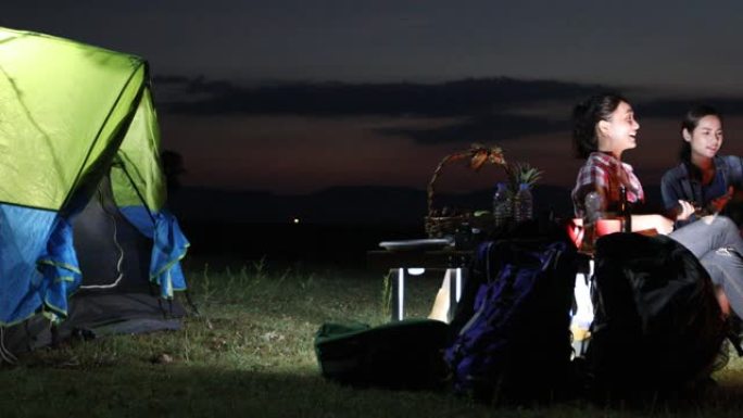 一群亚洲朋友的游客正在玩Ukelele，而他的放松的朋友正在一起坐在帐篷附近的篝火中聆听和扔柴火