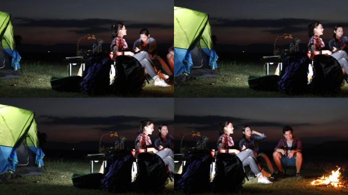 一群亚洲朋友的游客正在玩Ukelele，而他的放松的朋友正在一起坐在帐篷附近的篝火中聆听和扔柴火