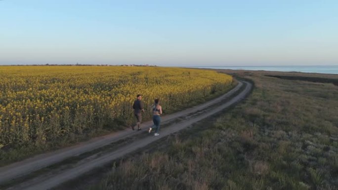 户外运动，女运动员和运动员在大自然有氧运动期间沿着油菜籽盛开的田野附近的道路奔跑