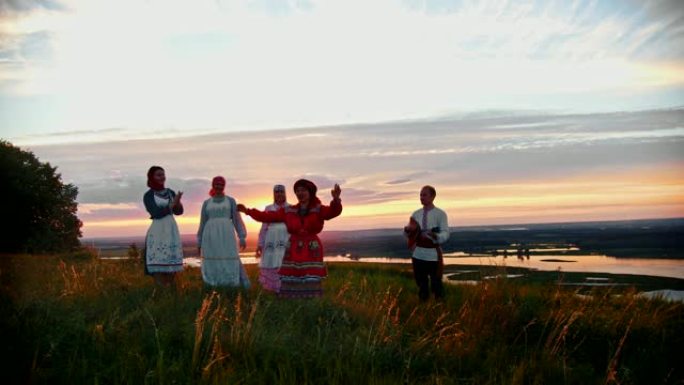 穿着俄罗斯传统服装的年轻人在美丽的日落背景下在球场上玩耍-一个男人在演奏balalaika和女人在音