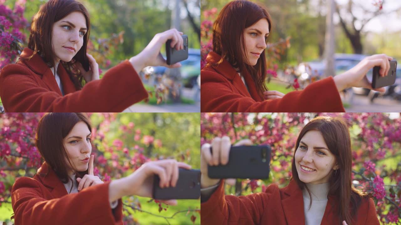 年轻迷人的红发女人在公园日落时在智能手机上的樱桃花或樱花花春天的背景上自拍