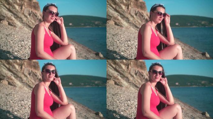 穿着红色连体泳衣和眼镜的美丽怀孕女孩坐在海滩上。