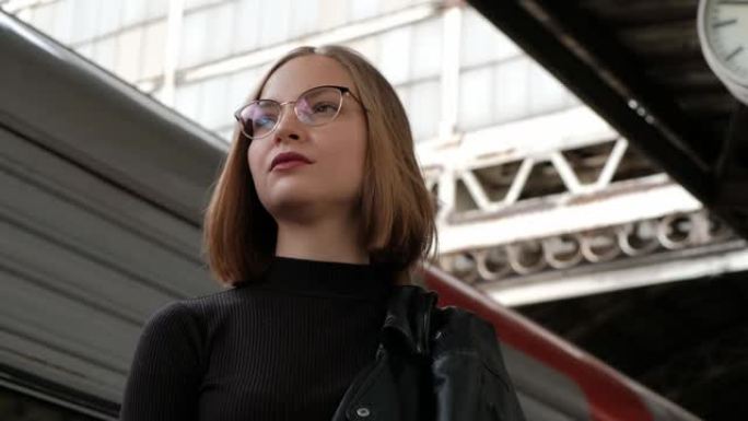 迷人的红发年轻女子短发，戴着眼镜，穿着黑色皮夹克，站在圣彼得堡美丽的历史悠久的维捷布斯克火车站，等待