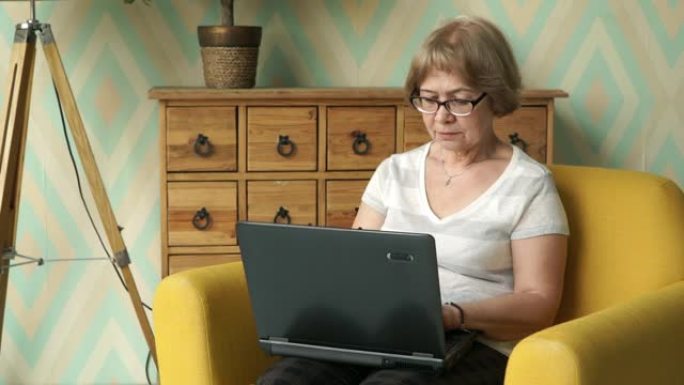 严肃的上了年纪的漂亮白种女人戴着眼镜坐在家里的椅子上用笔记本电脑工作。