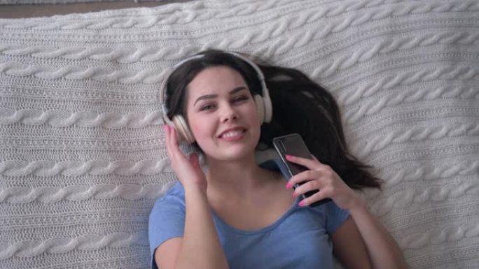 可爱的微笑女孩戴着智能手机听歌，一边在家床上放松，周末特写镜头，顶视图