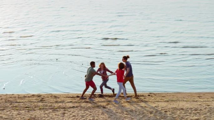 四个快乐的多民族朋友在海边的海滩上手牵手跳舞。年轻人在夏日从宽阔的镜头上感到快乐和无忧无虑