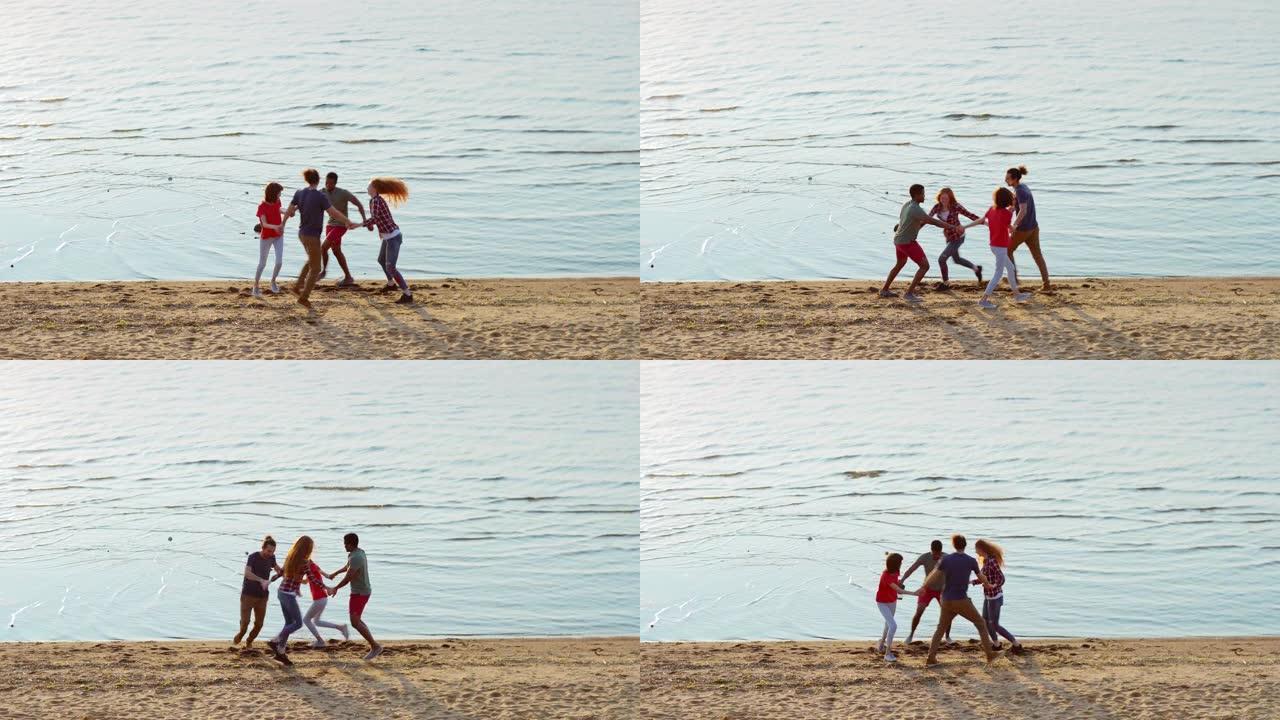 四个快乐的多民族朋友在海边的海滩上手牵手跳舞。年轻人在夏日从宽阔的镜头上感到快乐和无忧无虑