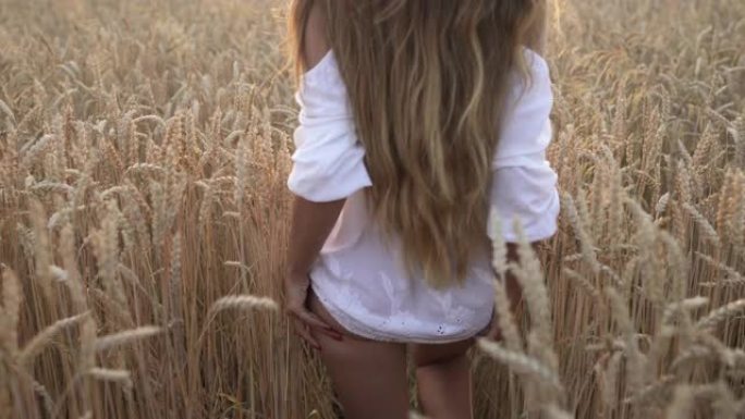 日落时分，穿着休闲风格白色连衣裙的年轻女子在麦田里散步的特写镜头。阳光落在金色无尽的田野上。