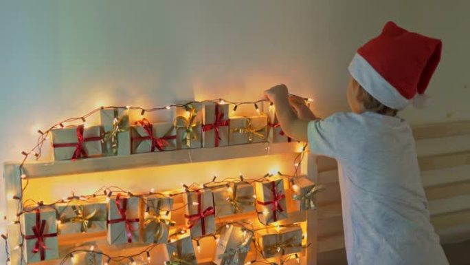 小男孩从挂在用圣诞灯照亮的床上的降临日历上拿了一份礼物。为圣诞节和新年概念做准备。降临日历概念