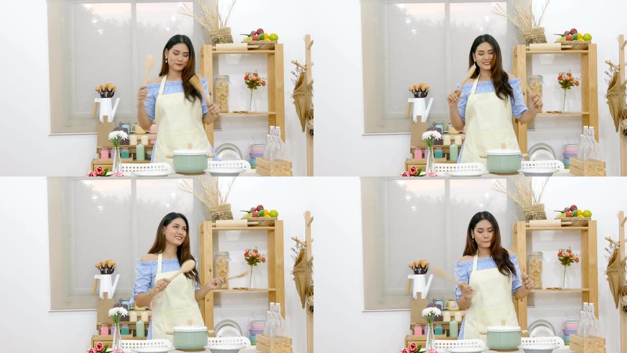 4K.美丽的亚洲女人拿着钢包在厨房里跳舞，喜欢在餐桌后面用锅，盘子和带有笑脸的厨具做饭