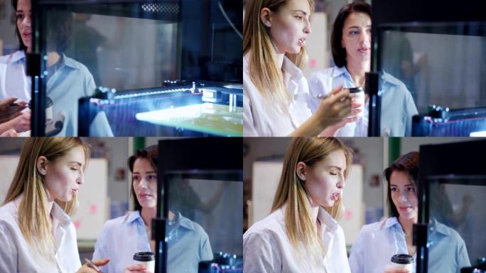 两名女工程师站在实验室的3d打印机旁，检查打印过程。女人向拿着咖啡杯的同事解释细节，特写镜头跟踪左镜