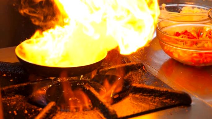 在餐厅的现代厨房里，无法识别的厨师在平底锅上烹饪flambe风格的菜肴。专业烹饪在燃气板上燃烧的煎锅