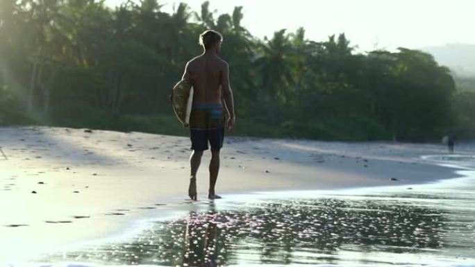 慢动作。金发肌肉冲浪者手持冲浪板走在海滩上，日出之光