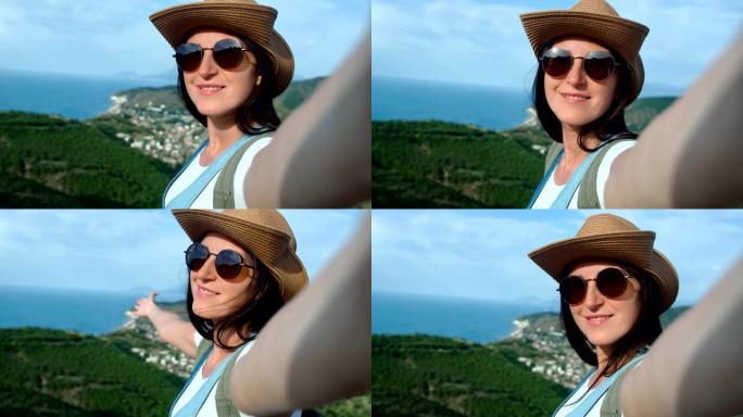 戴着墨镜和帽子的特写微笑旅行女人自拍，展示海景享受假期