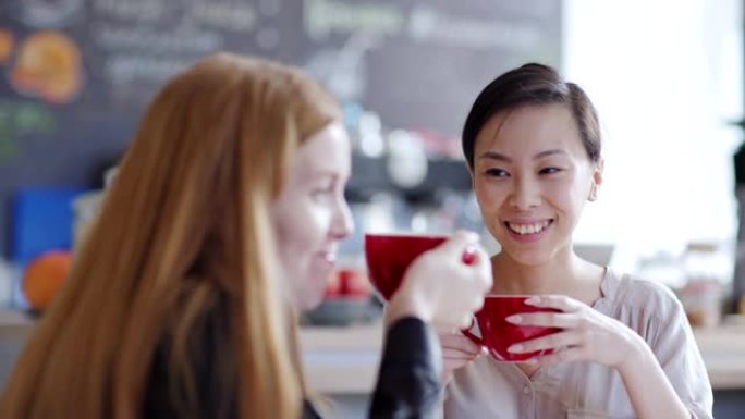 追踪两名年轻女子在咖啡馆喝茶或喝咖啡并聊天的正确镜头。亚洲女性向散焦的高加索朋友讲故事